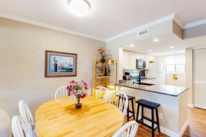A dining area with a wooden table and white chairs, a kitchen with white cabinets, and a small island with a sink. - Fun & bright beach getaway with pool, easy beach access & pond view (Hilton Head)
