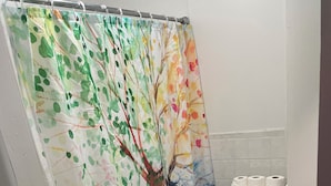 A bathroom with a shower curtain featuring a tree design, a toilet, a sink, and two mats. -  1-bedroom Temple near train and buses (Philadelphia)