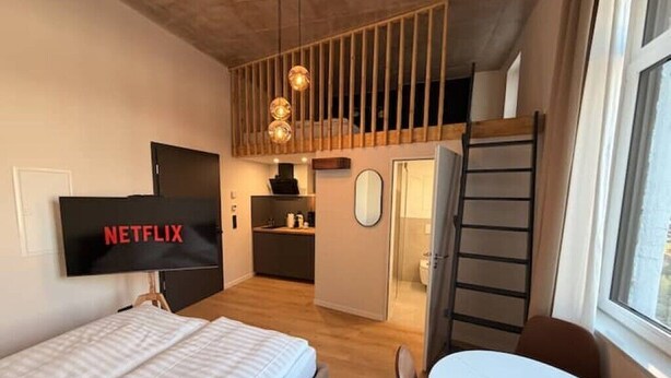 Une chambre d’hôtel moderne équipée d’un lit, d’un téléviseur diffusant « NETFLIX », d’une table à manger, d’un coin cuisine et d’un espace en mezzanine accessible par une échelle.