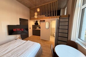A modern hotel room with a bed, a TV displaying 'NETFLIX,' a dining table, a kitchen area, and a loft space with a ladder. - Villa Weitmar: The urban studio (Bochum)