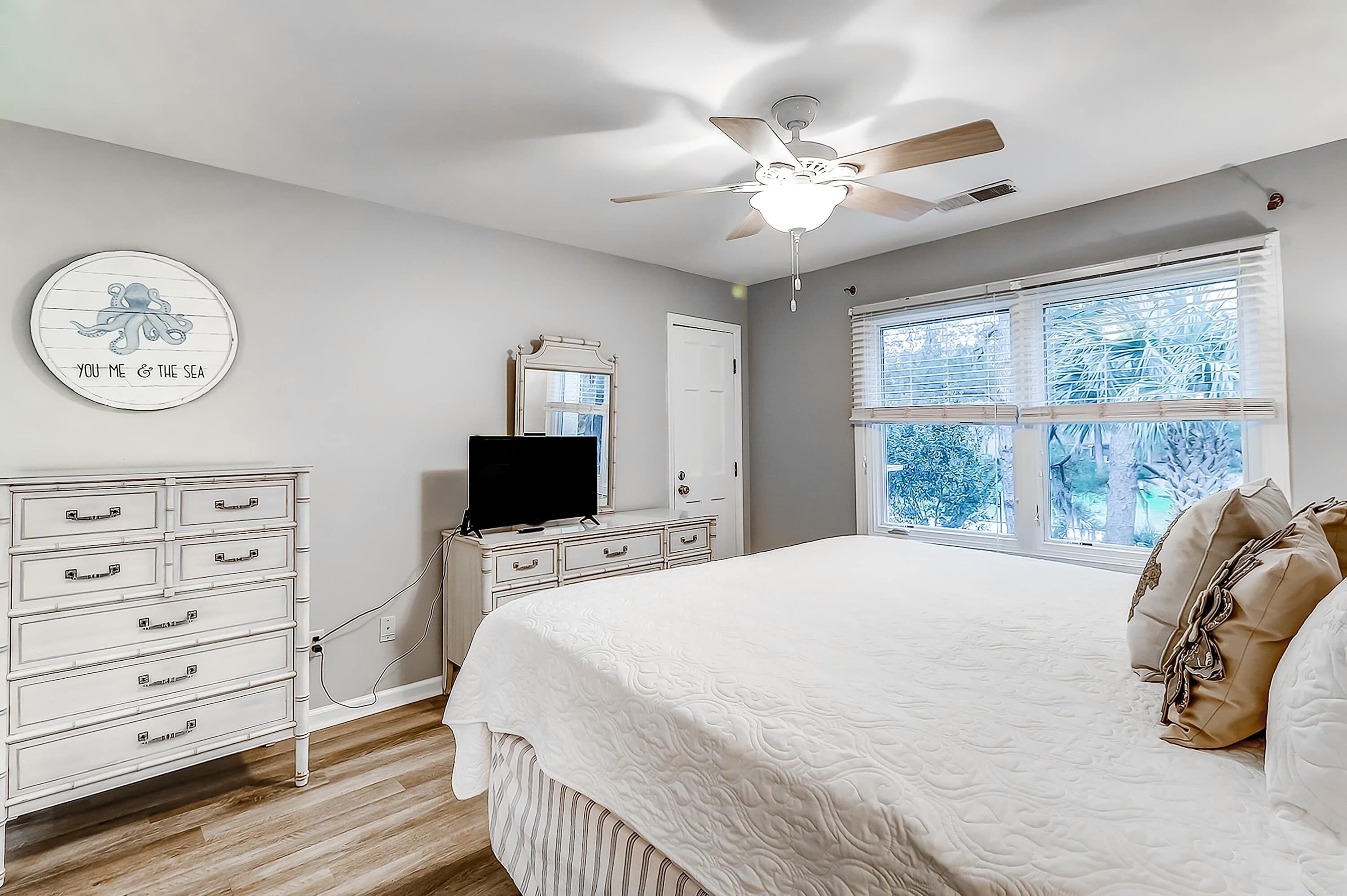 A bedroom with a bed, dresser, TV, ceiling fan, and a window with blinds.
