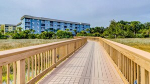 En træpromenade fører hen til en blå bygning med flere balkoner, omgivet af grøn bevoksning.
