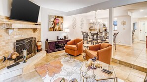 A living room with a fireplace, a flat-screen TV, orange armchairs, a coffee table, and a dining area with chairs and a chandelier. - Dog Friendly Villa with Walkable Beach Access, AC, Deck, WiFi & Shared Pool (Hilton Head)