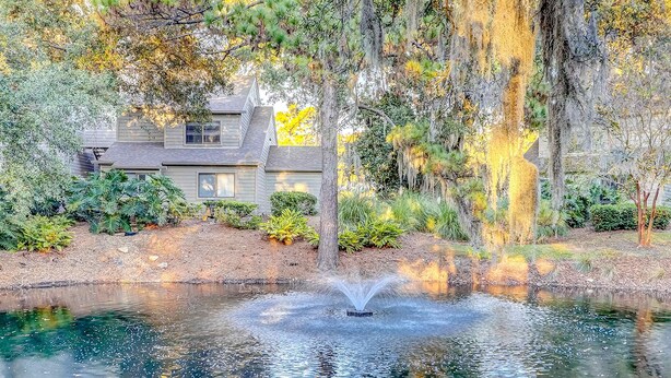 A pond with a fountain, surrounded by trees and a house. - Snowbird Friendly villa with Golf View, Patio, Shared Pool, Tennis, AC, WiFi (Hilton Head)