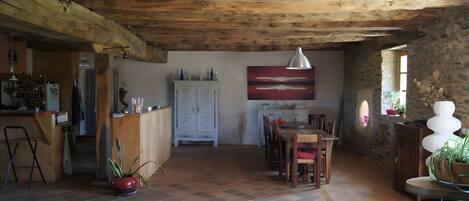 A rustic interior with a wooden ceiling, a dining area with a table and chairs, a kitchen with a refrigerator, and a large abstract painting on the wall.
