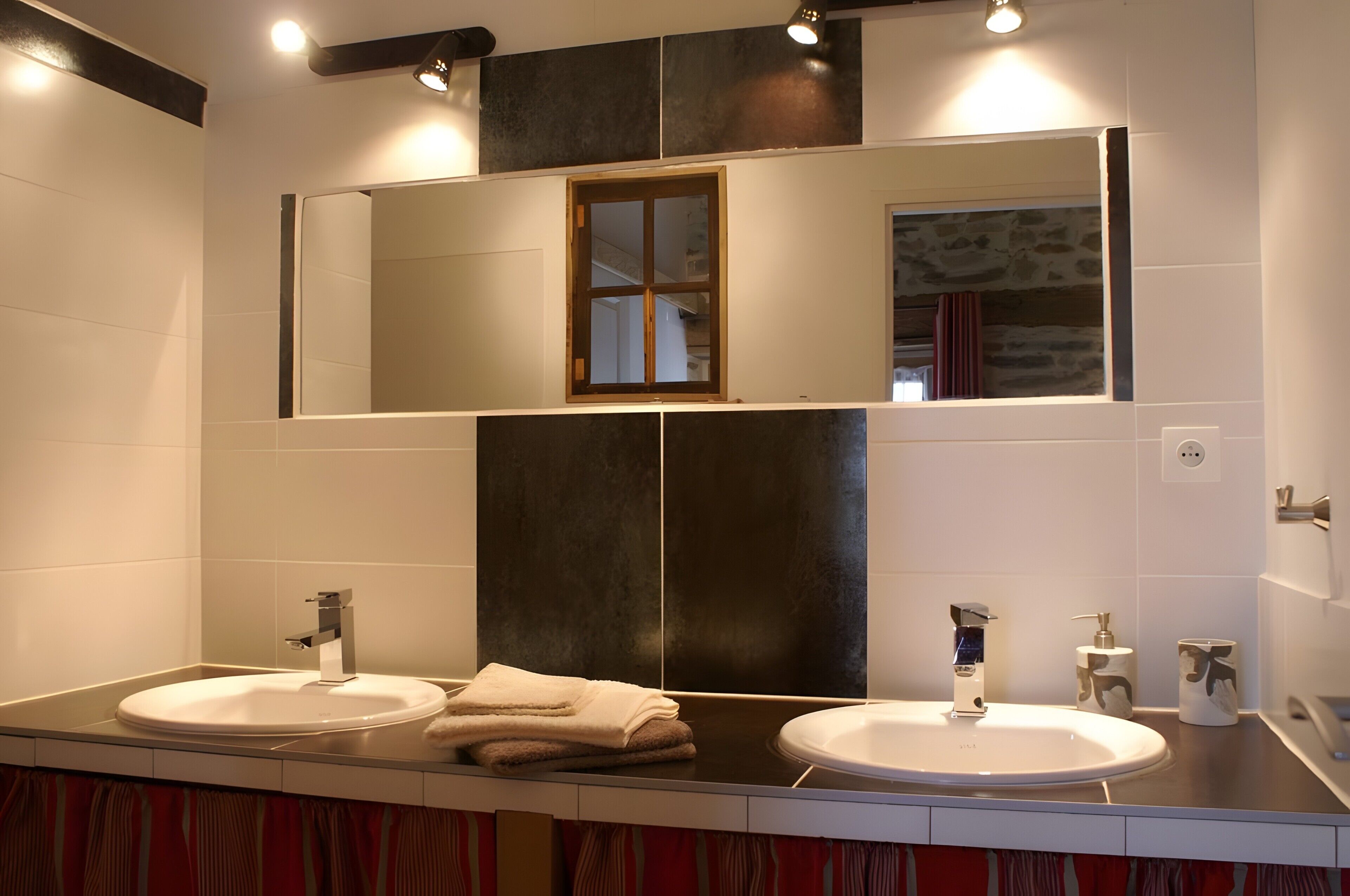 Une salle de bain avec deux lavabos, un grand miroir et une fenêtre donnant sur un mur en pierre.