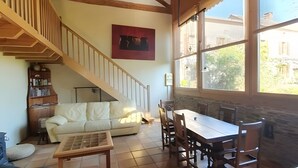 A spacious living area with a wooden staircase, a dining table with chairs, a sofa, and a large window providing a view of the outside.
