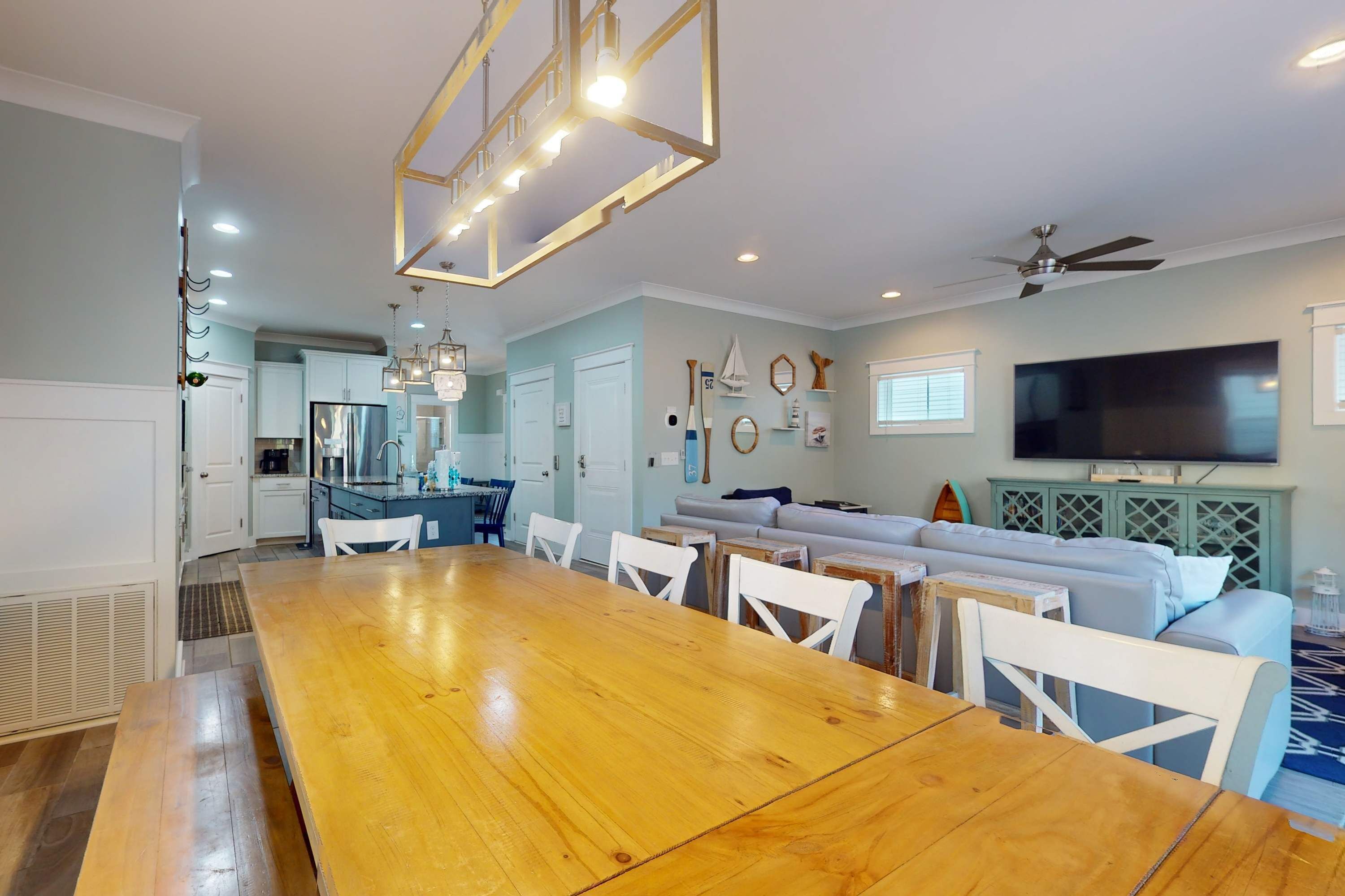 A spacious dining area with a wooden table and white chairs, a kitchen with white cabinets, a living area with a sofa and TV, and a ceiling fan.