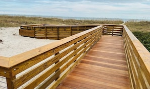 Ein hölzerner Steg führt zu einem Strand mit Sand und Dünen im Hintergrund.