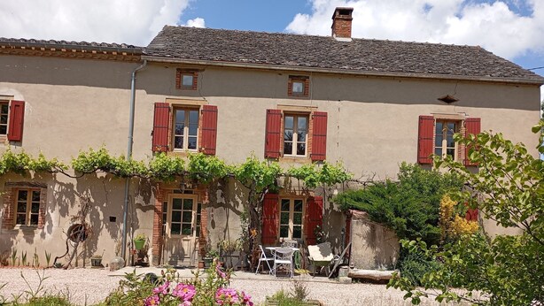 Un bâtiment à deux étages avec des volets rouges, un toit en tuiles et un jardin agrémenté de mobilier d’extérieur.