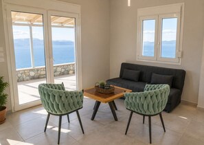 A room with a sofa, two chairs, a table, and a view of the sea through a glass door. - Nireos Coast Evia - Panoramic Seaview Gem (Akti Nireos)