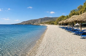 A pebble beach with clear water, a row of blue lounge chairs, and a mountain in the background.