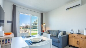 A modern living room with a dining area, a grey sofa, a wooden cabinet, and a view of a building through the window. - Affittimoderni Castelsardo - White Breeze (Castelsardo)