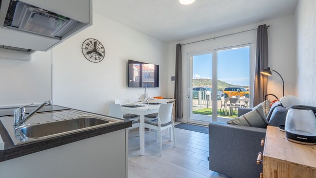 A modern kitchen with a built-in sink, dining area with a table and chairs, a gray sofa, and a large sliding glass door leading to an outdoor patio. - Affittimoderni Castelsardo - White Breeze (Castelsardo)