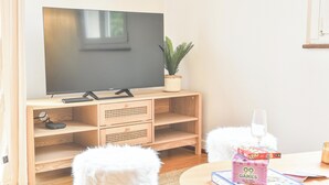 A room with a television on a wooden stand, a small plant, a fluffy white stool, and a table with board games. - Rock in Share - Bianca – 2-Bedroom Apartment with Parking Included & Tram (Strasbourg)