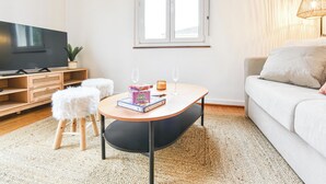 A living room with a flat-screen TV, a small round table with a glass, a sofa, and a window with blinds. - Rock in Share - Bianca – 2-Bedroom Apartment with Parking Included & Tram (Strasbourg)