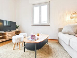 Ett vardagsrum med en platt-TV, ett litet runt bord med ett glas på, en soffa och ett fönster med persienner.