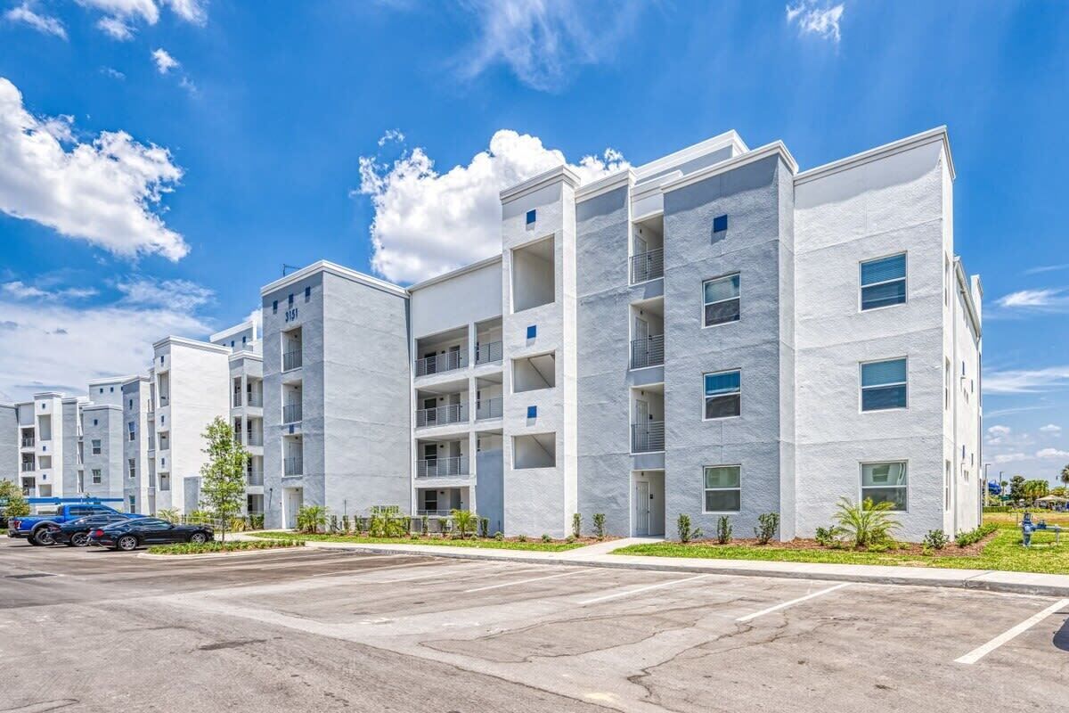 A multi-story residential building with balconies, a parking lot, and a clear sky.