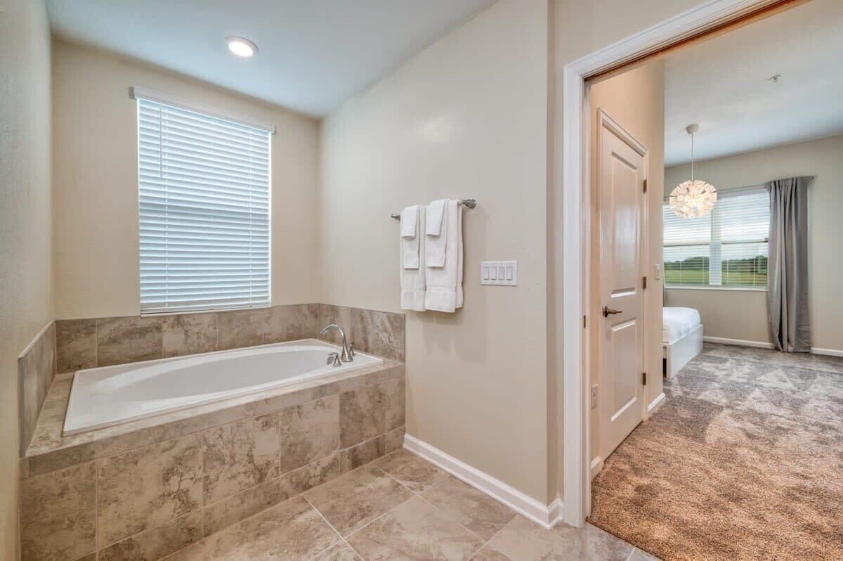 A bathroom with a bathtub, a window with blinds, a towel rack, and a door leading to another room.