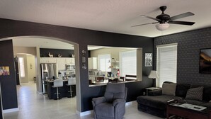 A modern living room with a grey armchair, a dark sofa, and a coffee table. A kitchen with white cabinets and a dining area with a table and chairs is visible in the background. - Little Peace and Quiet out on Little Leaf (San Tan Valley)