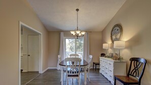 A dining area with a chandelier, a white dining table, and chairs. There is a window with curtains, a white cabinet with drawers, and a wall-mounted lamp.