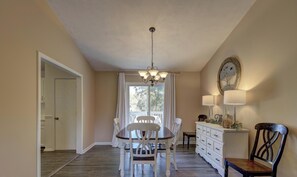 Un comedor con una araña, una mesa blanca y sillas. Hay una ventana con cortinas, un armario blanco con cajones y una lámpara de pared.