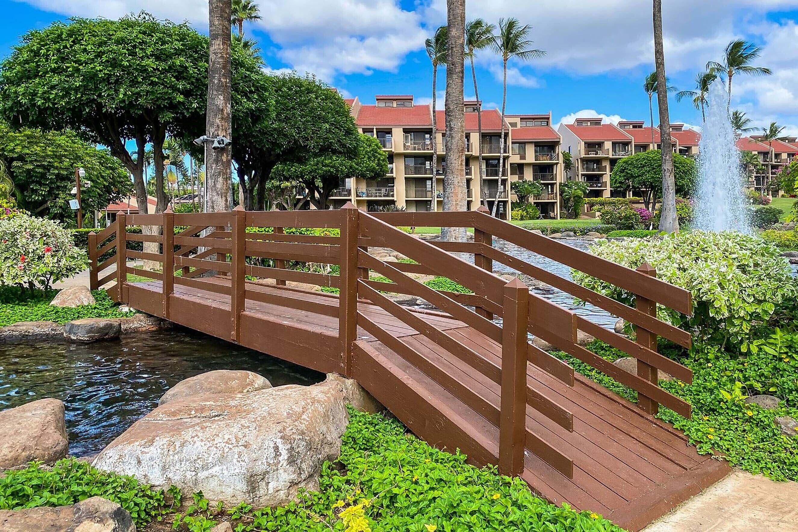 A wooden bridge over a stream, surrounded by lush greenery and palm trees, with a hotel building in the background.
