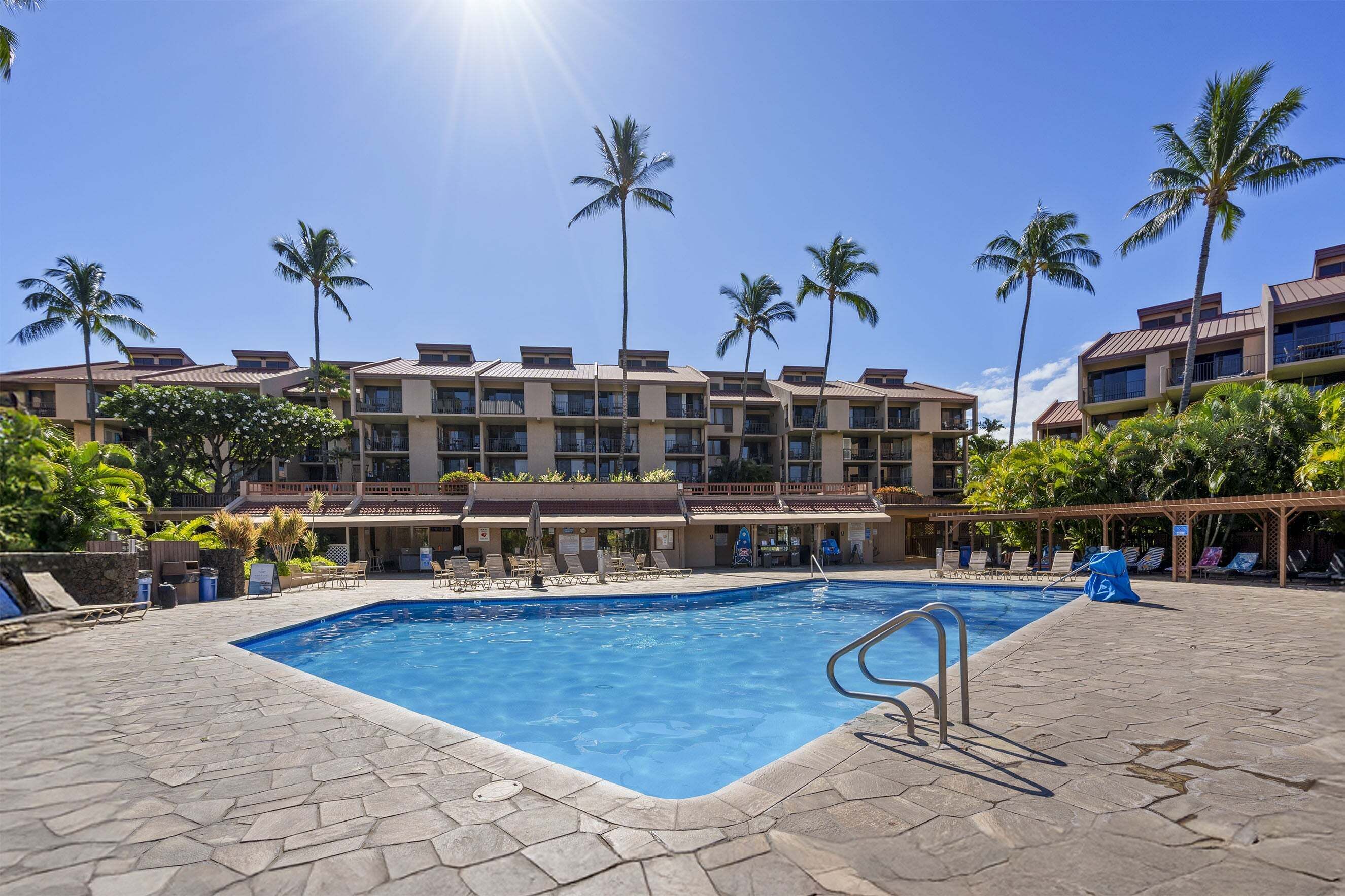 Ein Swimmingpool mit klarem, blauem Wasser, umgeben von Liegestühlen und Palmen, mit einem Hotel im Hintergrund.