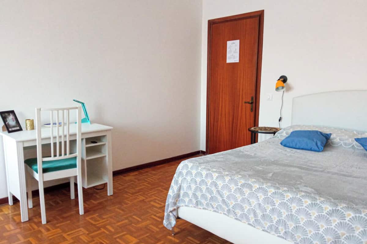 Un dormitorio con cama, escritorio y silla, puerta de madera y una pequeña mesa con lámpara.