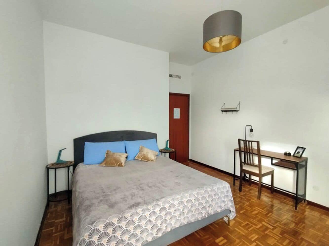 Un dormitorio con cama, escritorio, silla, lámpara y puerta.