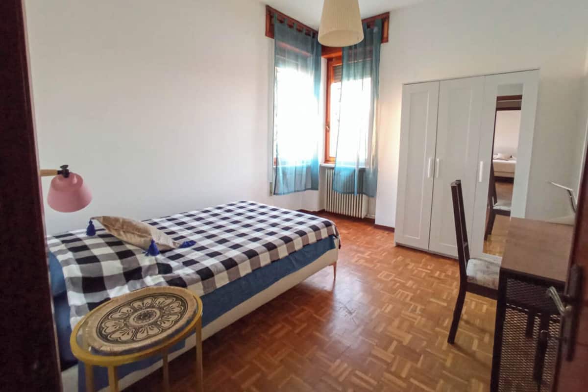 Un dormitorio con una colcha a cuadros, una mesita redonda pequeña, una lámpara colgante, un armario y una ventana con cortinas.