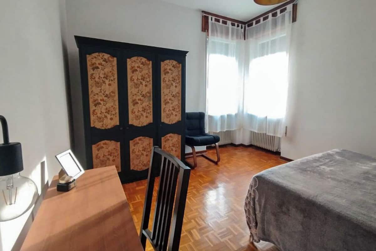 Una habitación con suelo de madera, una cama, una silla, un escritorio con lámpara y una ventana con cortinas.