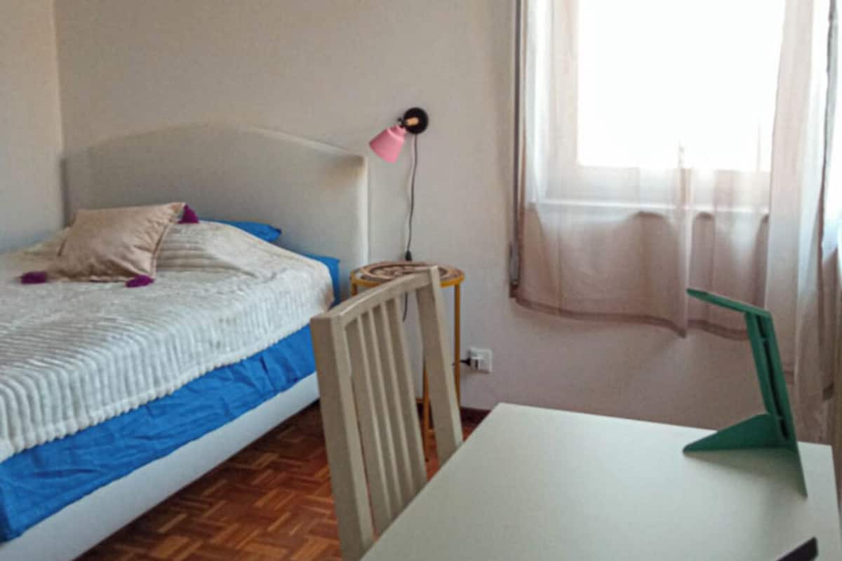 Un dormitorio con cama, escritorio, silla, lámpara y ventana con cortinas.