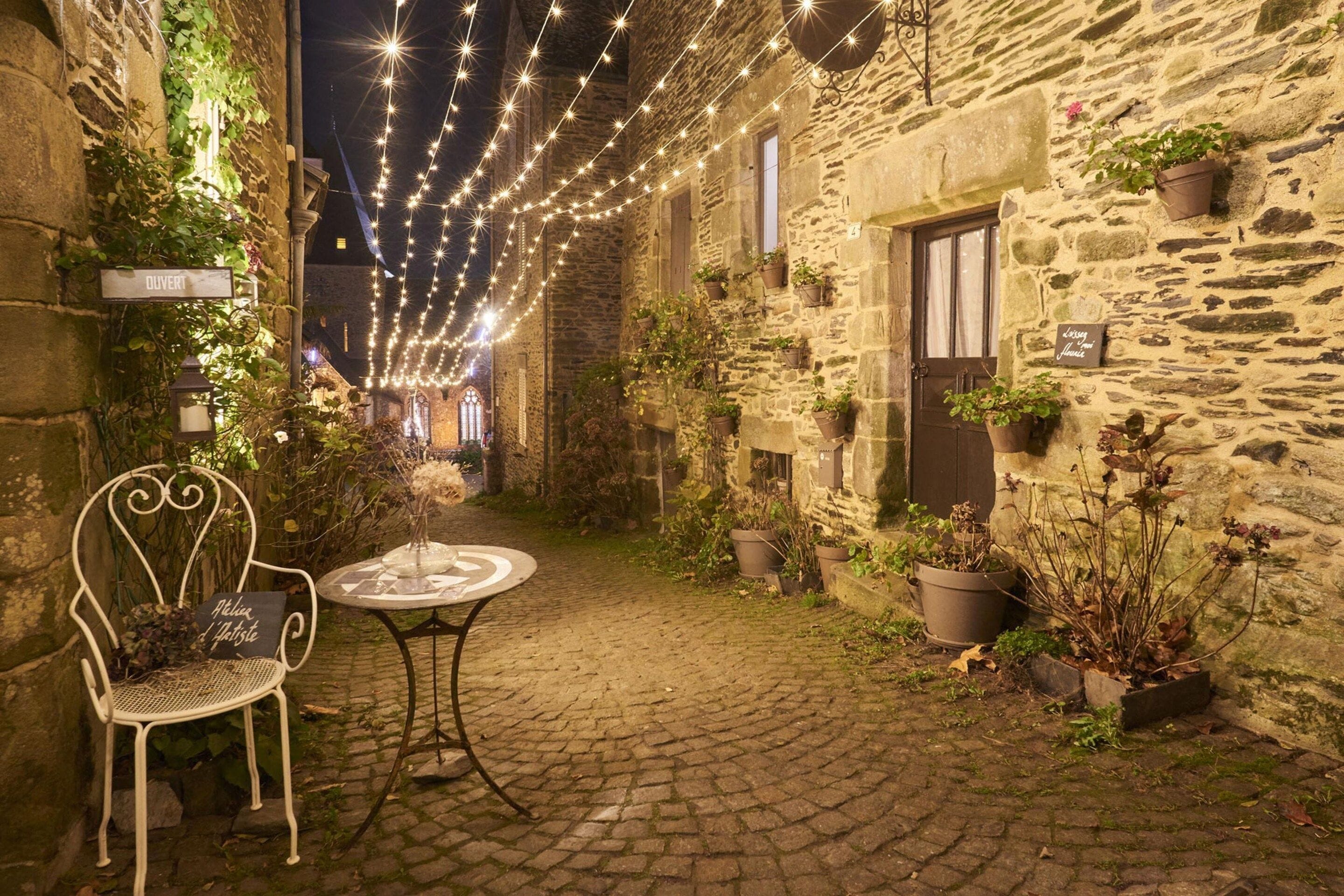 Un edificio de piedra con un patio adoquinado, una silla de metal blanco, una mesa redonda y luces colgantes.