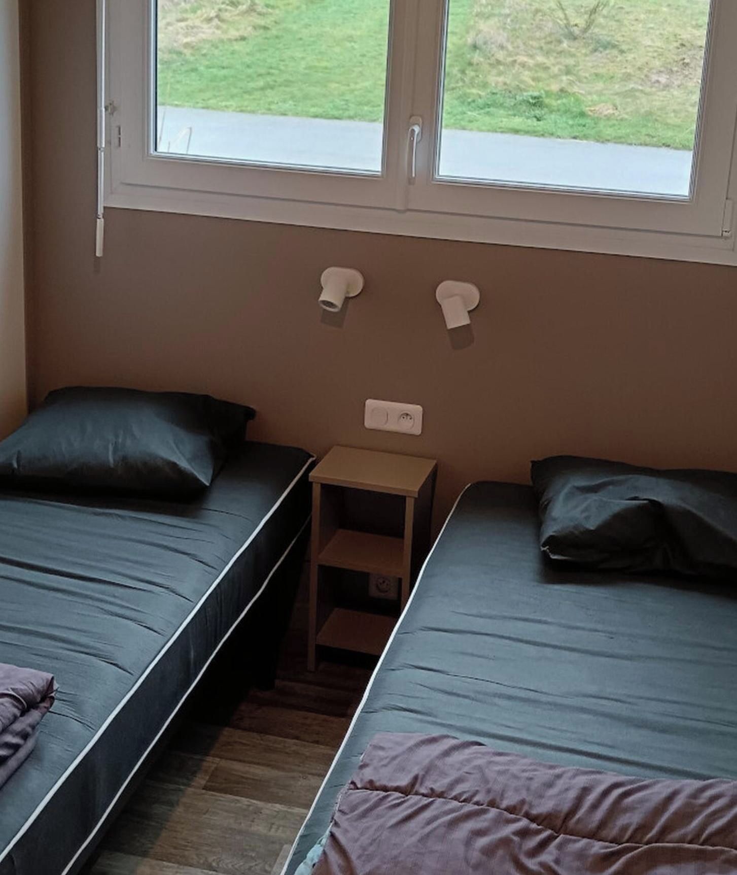 A room with two single beds, a small wooden nightstand, and a window with a view of the outdoors.