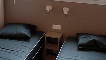 A room with two single beds, a small wooden nightstand, and a window with a view of the outdoors.
