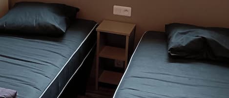 A room with two single beds, a small wooden nightstand, and a window with a view of the outdoors.