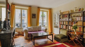 A cozy living room with a sofa, a coffee table, a bookshelf, and a view of the city through large windows. - Paris Center St Claude residence (Paris)