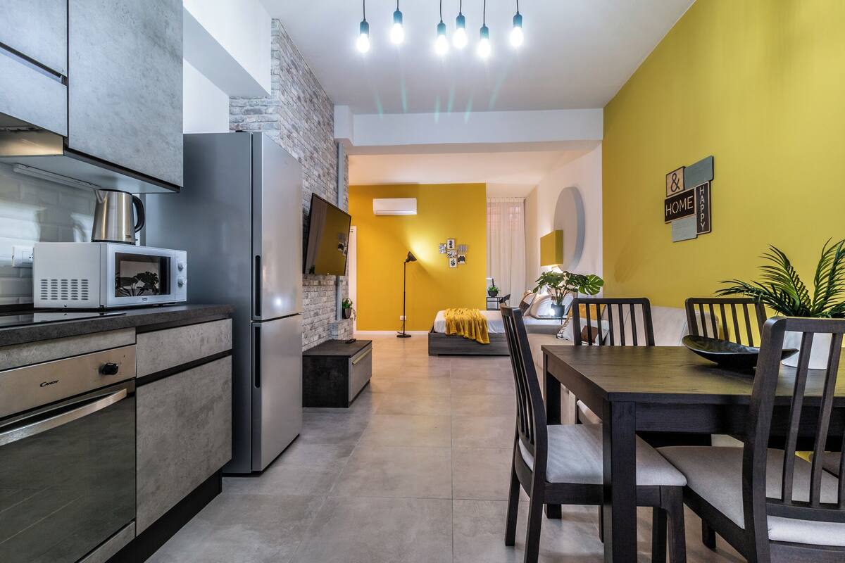 A modern kitchen with a refrigerator, microwave, and kettle, a dining area with a table and chairs, and a yellow accent wall.