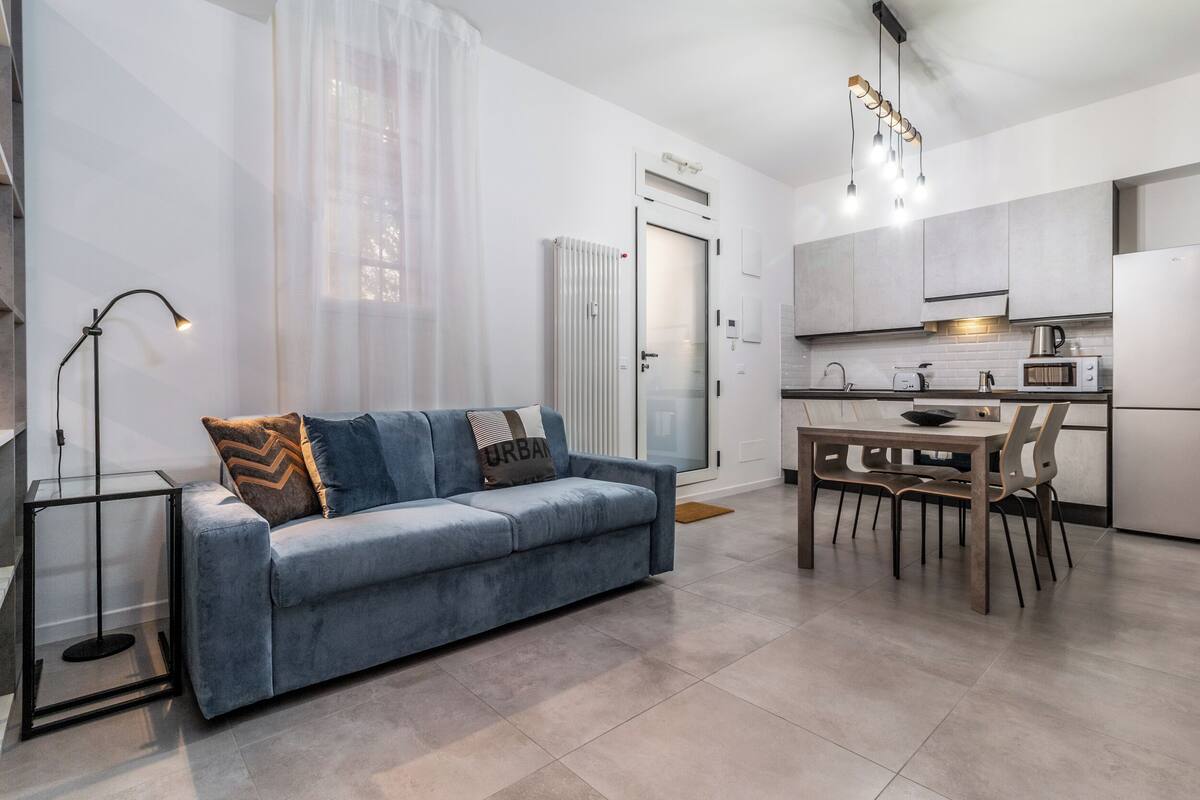 A modern living room with a sofa, a dining table with chairs, a kitchen area with cabinets and appliances, and a radiator.