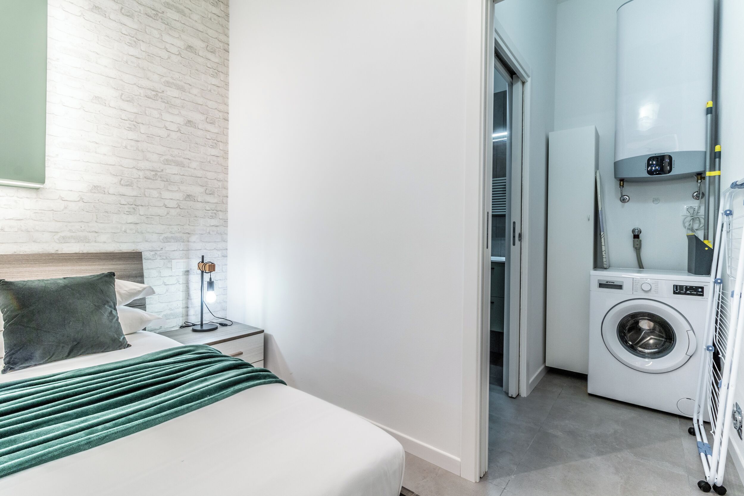 A hotel room with a bed, a green throw, a nightstand, a lamp, a washing machine, and a mirror.
