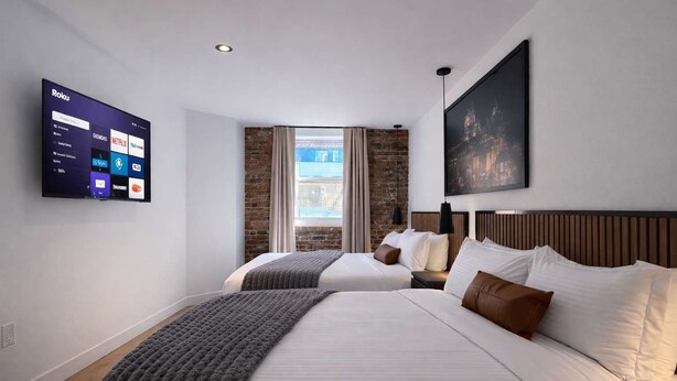 A hotel room with a large bed, a smaller bed, a TV mounted on the wall, and a window with a view of buildings. - Room with 2 Queen Beds | Downtown Quebec City (Québec City)