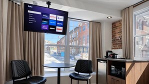 Ein modernes Zimmer mit Fernseher, der Streaming-Dienste anzeigt, zwei schwarzen Stühlen, einem kleinen Tisch, einer Küchenzeile und Blick auf die Straße.