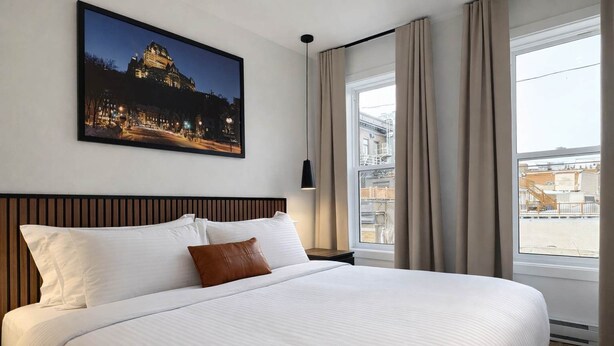 Habitación de hotel con una cama grande, cabecera, mesita de noche, lámpara, ventana con cortinas y vista a la ciudad.