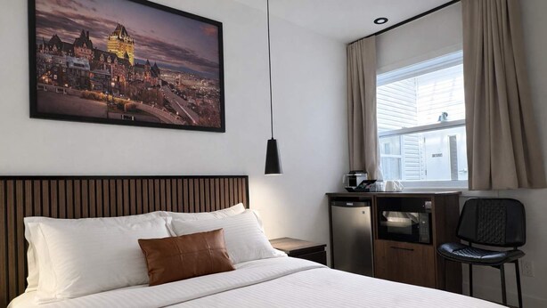 Een hotelkamer met een bed, een bureau met een koelkast, een stoel, een raam met gordijnen en een stadsgezichtschilderij aan de muur.