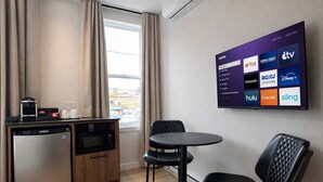 Camera d'albergo compatta con angolo cottura, zona pranzo con tavolo e sedie, e televisore a parete che mostra servizi di streaming.