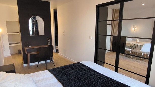 A bedroom with a large bed, a desk, a chair, a mirror, and a sliding glass door leading to a dining area. - Sublime central T2 (Montélimar)