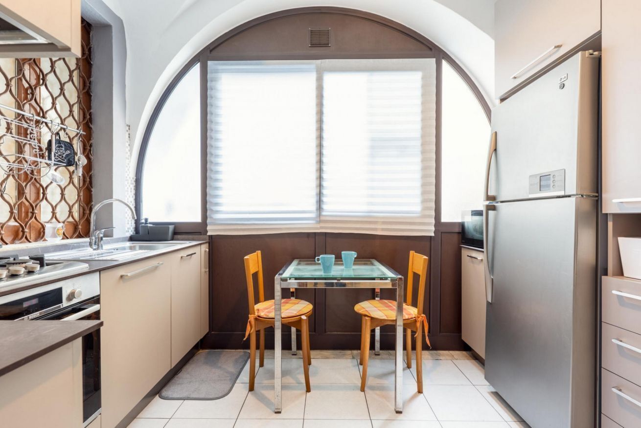 A compact kitchen with a dining table, two chairs, a refrigerator, and a window with blinds.