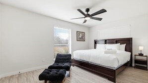 Quarto com cama, ventilador de teto, janela com persianas, mesa de cabeceira e uma cadeira.