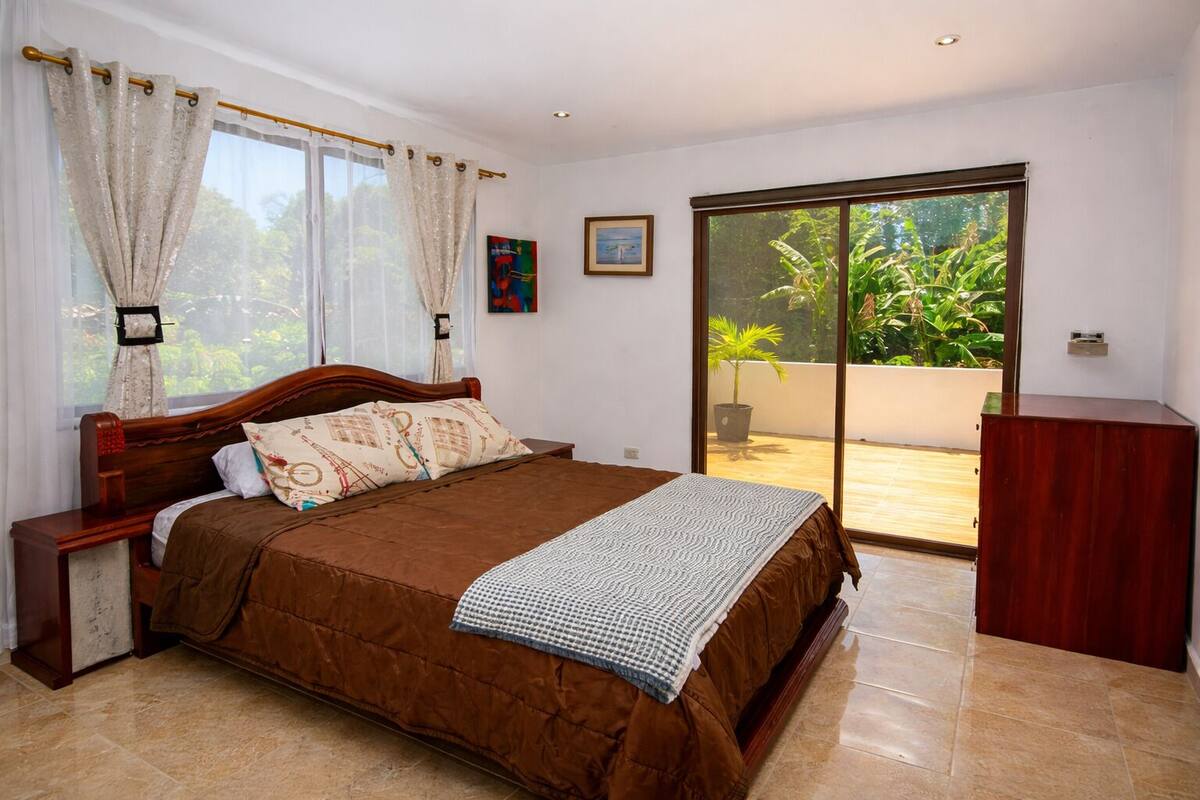 A bedroom with a bed, curtains, a painting, a window, and a sliding door to a balcony.